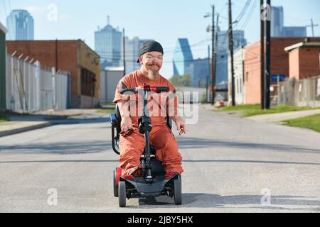 Ritratto di uomo adulto in strada in Texas su uno scooter per la mobilità che guarda in macchina fotografica con lo skyline di Dallas sullo sfondo. Foto Stock