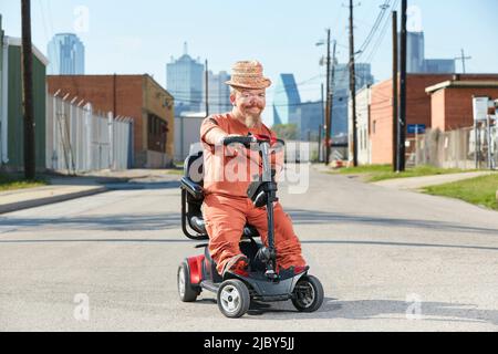 Ritratto di maschio caucasico adulto piccola persona su uno scooter di mobilità parcheggiato in mezzo alla strada con Dallas Texas skyline sullo sfondo. Foto Stock