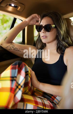 Ritratto di donna vestita casualmente in occhiali da sole con tatuaggi seduti sul sedile posteriore di berlina di lusso Foto Stock