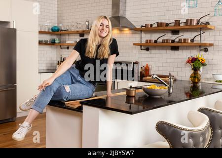 Ritratto di sorridente giovane donna caucasica guardando la macchina fotografica sorridente, mentre seduto sul piano di lavoro della sua cucina appartamento. Foto Stock