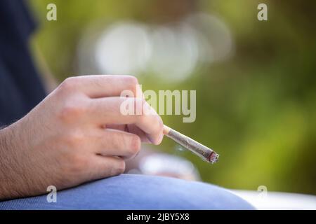 Dettaglio dell'uomo che tiene un giunto di pre-roll del CBD illuminato, sigaretta con fumo che sale all'aperto Foto Stock