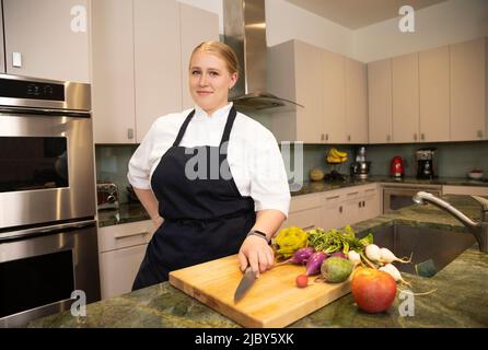 Chef Megan Gill in una cucina guardando la macchina fotografica con fiducia, mentre avendo una mano sul suo coltello da cucina. Foto Stock