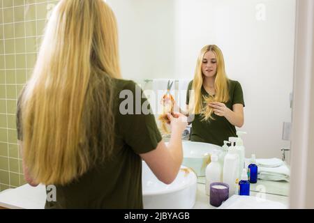 Giovane donna in bagno di casa guardando la ciocca di capelli dopo aver tagliato i propri capelli con le forbici Foto Stock