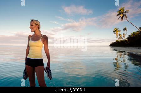 Giovane donna in piedi sulla spiaggia tropicale tenendo le scarpe e sorridendo Foto Stock
