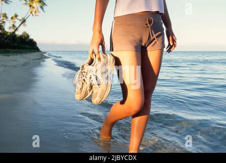 Giovane donna che cammina lungo la spiaggia tropicale tenendo le sue scarpe Foto Stock