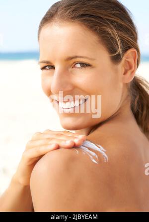 Ritratto di sana bella giovane donna che applica la crema solare alla spalla alla spiaggia e sorridente Foto Stock