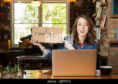 Ritratto di giovane donna seduta al tavolo in libreria caffetteria con computer portatile, tenendo a mano segno fatto che legge ÒI hanno idee Foto Stock