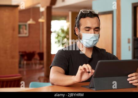 Giovane uomo ispanico che indossa maglietta nera e occhiali trasparenti con maschera facciale, seduto al bar in cucina del centro loft con iPad Foto Stock