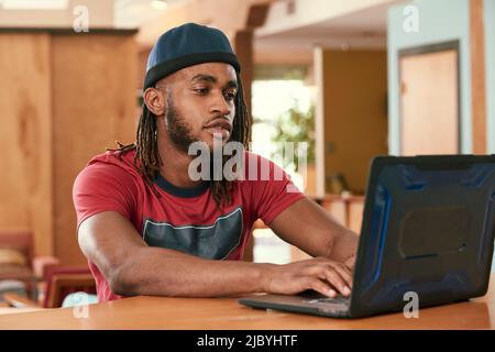 Ritratto di giovane uomo etnico con maglietta rossa e cappello a maglia , seduto al bar in cucina del centro loft con computer portatile Foto Stock