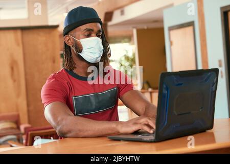 Ritratto di giovane uomo etnico con maglietta rossa e cappello a maglia con maschera facciale, seduto al bar in cucina del centro loft con computer portatile Foto Stock