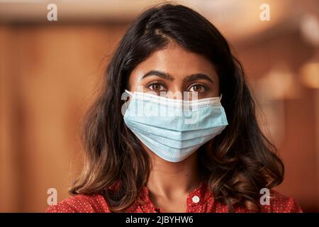 Ritratto di giovane etnia donna indossare maschera viso indossare camicetta arancione seduta al bar in cucina del centro loft Foto Stock