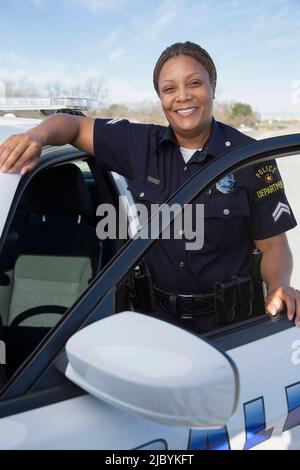 Poliziotto in piedi alla porta della macchina di polizia guardando verso la macchina fotografica sorridente Foto Stock