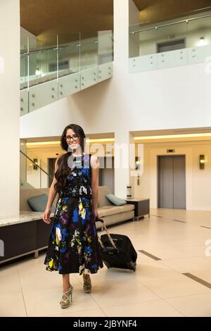 Donna ispanica tirando valigia scorrevole nella hall hotel Foto Stock