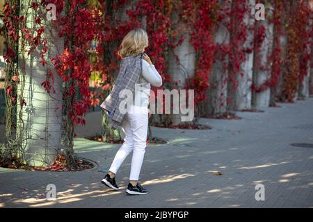 Elegante bionda 35-40 anni in pantaloni bianchi e giacca passeggiate a piedi attraverso la città autunnale decorata con verde rosso brillante di edera soprastante Foto Stock