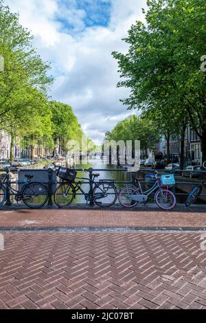 Biciclette parcheggiate su un pavimento piastrellato ponte sul canale, ormeggiate barche sull'acqua. Amsterdam, Paesi Bassi. Case tradizionali facciata in mattoni e alberi Foto Stock