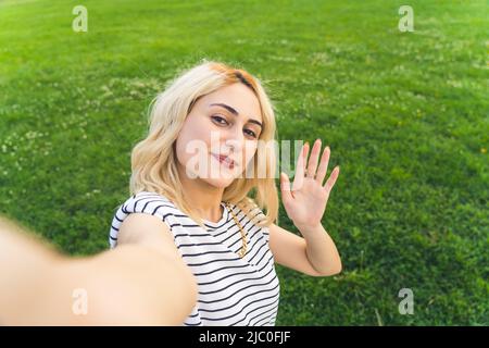 foto di una giovane ragazza allegra bionda in piedi nel parco e prendere un selfie ondeggiare medio primo piano all'aperto. Foto di alta qualità Foto Stock