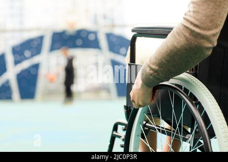 Primo piano di un inriconoscibile uomo disabile in maglione marrone che gira su sedia a rotelle manuale mentre guarda il giocatore di basket Foto Stock