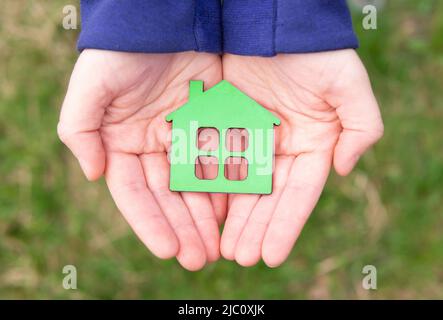 Vista dall'alto di un modello di casa in legno dipinto di verde chiaro posto in entrambe le mani all'aperto. Concetto di offerta di Dream House. Foto Stock