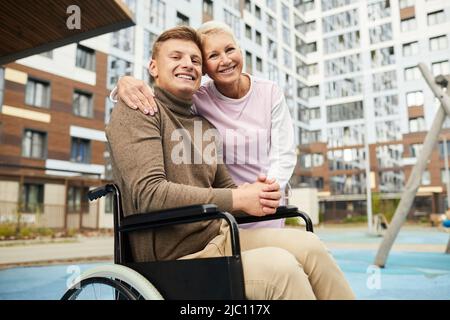 Ritratto di tipo positivo infermiera qualificata che abbraccia l'uomo in sedia a rotelle mentre camminano sopra il cortile vicino agli edifici di appartamento Foto Stock