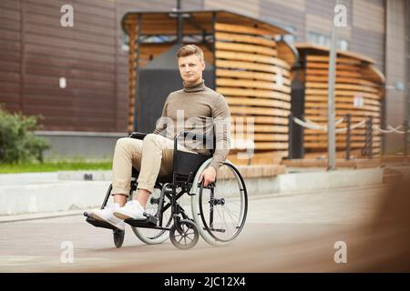 Contenuto bel ragazzo giovane con lesione del midollo spinale seduta in sedia a rotelle all'aperto e guardando la macchina fotografica Foto Stock