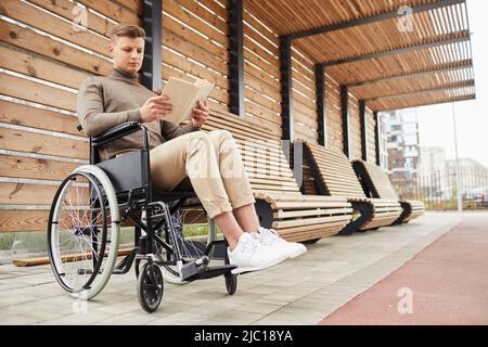 Giovane handicappato serio in scarpe bianche seduto in sedia a rotelle alla stazione ferroviaria e libro di lettura Foto Stock