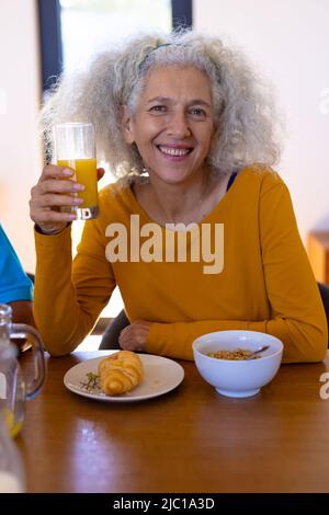 Ritratto di donna anziana caucasica con croissant, avena e succo di frutta a colazione al tavolo da pranzo Foto Stock
