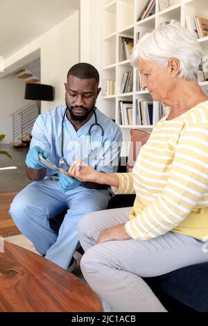 Operatore sanitario africano americano maschile che misura lo zucchero nel sangue della donna anziana caucasica utilizzando glucometro Foto Stock