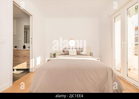 Camera da letto con letto matrimoniale con copriletto grigio, mobili bianchi e testiera, balcone con porte in alluminio e pavimento in legno chiaro Foto Stock