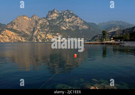 Tranquilla mattina presto vicino alla punta settentrionale del Lago di Garda in Trentino-Alto Adige, Italia, con una linea di boe di corda che conduce attraverso l'acqua placida verso il porto di Torbole sul Garda, un ex posto di frontiera veneziano e austriaco oggi popolare tra marinai, kiteboarder e windsurf. Foto Stock