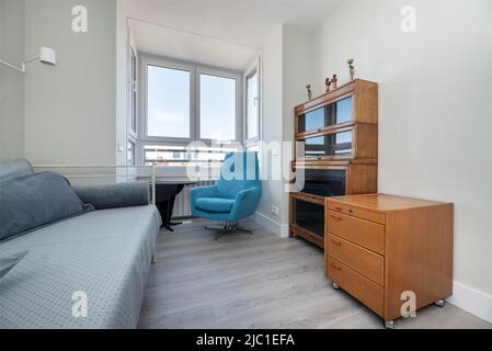 Angolo di camera con divano singolo blu, finestra con vista, divano-letto con telaio in metallo bianco, pavimento in parquet grigio e libreria di design con Foto Stock