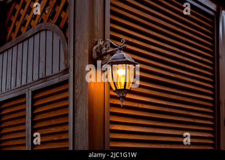 lanterna retrò forgiata in ferro con una paralume di vetro e una lampadina elettrica su una parete di legno fatta di tavole, legno persiane architettura con lig sera Foto Stock