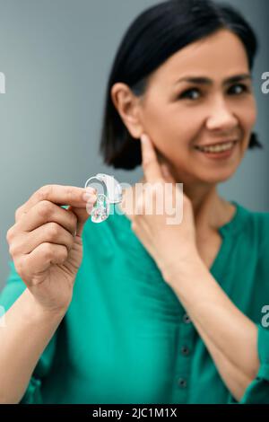 Trattamento della sordità in persone adulte. Donna che tiene l'apparecchio acustico BTE in mano in primo piano e punta verso l'orecchio Foto Stock