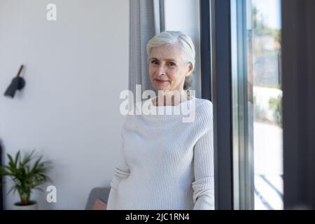 Ritratto di donna anziana caucasica sorridente in piedi vicino alla finestra a casa Foto Stock