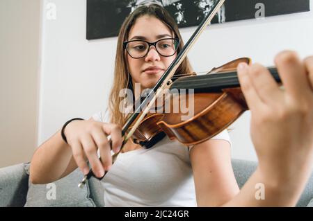 giovane latino caucasica venezuelana, indossando occhiali, vestito di bianco, concentrato a suonare il violino, esercitandosi seduto sul divano nel vivo r Foto Stock