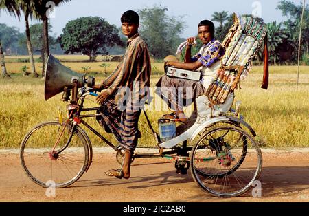 Bangladesh; uomo che annuncia l'arrivo di un circo in zona rurale mentre un ciclista lo guida in giro su un trisaw. Foto Stock