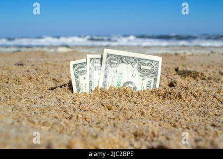 Carta fattura un dollaro sepolto nella sabbia sulla spiaggia sullo sfondo del mare primo piano. Banconote in denaro americane di un dollaro di denominazione in sabbia nella giornata estiva di sole. Concetto di finanza monetaria Foto Stock