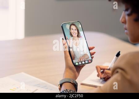 Donna d'affari biraciale che prende appunti mentre videochiama il collega caucasico femmina su smartphone Foto Stock