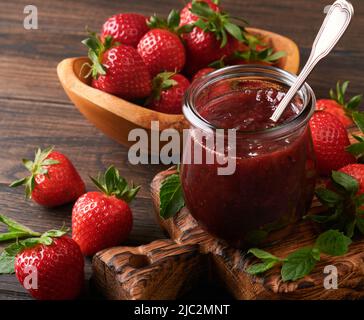 Marmellata di fragole. Marmellata di fragole in vaso di vetro con piatto di frutti di bosco freschi su un vecchio sfondo di legno scuro tavolo, primo piano. Marmellata di fragole fatte in casa. Foto Stock