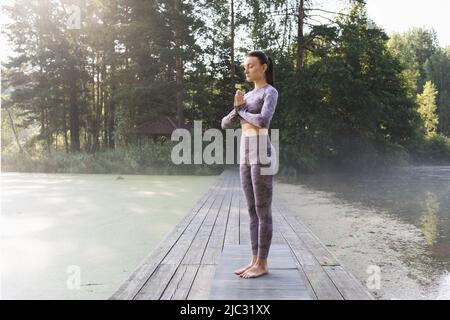 Una donna che pratica yoga, facendo una variazione dell'esercizio vrikshasana, albero posa con namaste, in piedi su un ponte di legno in una mattinata estiva nel Foto Stock