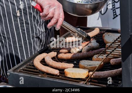 Salsicce di carne, pane, funghi vengono grigliati. Mano dell'uomo con pinze da cucina. Foto Stock