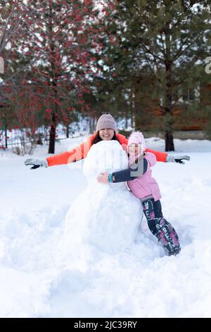 Donna con le mani semisulte in piedi sulla neve dietro un pupazzo di neve incompiuto con la bambina che lo abbraccia di sera con rowan e abeti sullo sfondo Foto Stock