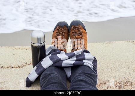 I piedi maschi in scarponi da trekking gialli sono avvolti in una sciarpa a righe e un thermos di tè caldo, caffè sullo sfondo dell'onda di mare in arrivo. IT Foto Stock