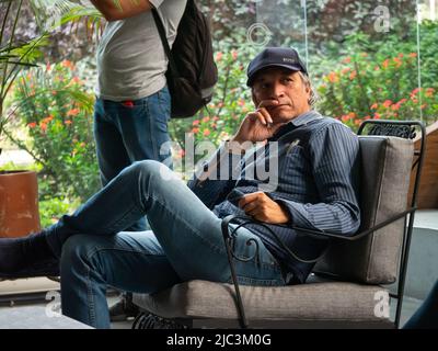 Medellin, Antioquia, Colombia - Febbraio 19 2022: Uomo dei capelli grigio sudamericano con un cappello nero fissa seduta in una sedia in un posto pubblico Foto Stock