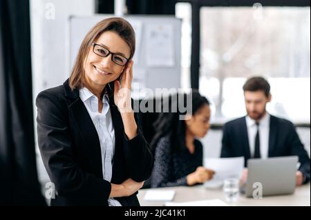 Positivo sicuro elegante splendida caucasica donna business con occhiali, in un abito formale, direttore creativo, business leader, in piedi in un ufficio moderno, guardando la macchina fotografica, sorridente amichevole Foto Stock