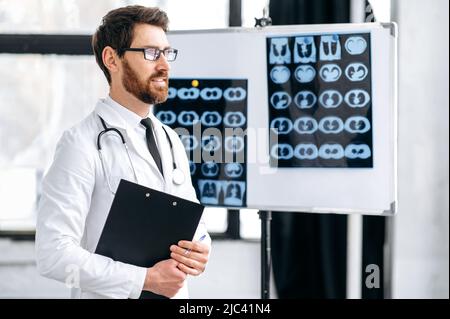 Orgoglioso pensivo caucasico maschio medicina generale medico, chirurgo, pulmonologista, in uniforme medica, con occhiali e stetoscopio sulle spalle, stare in clinica, guarda al lato, pensare Foto Stock
