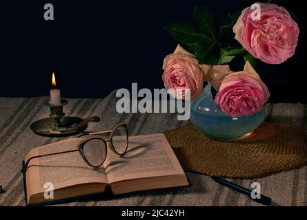 Inglese rosa in vaso su un tavolo accanto ad un libro aperto con bicchieri per la lettura e una candela bruciante con sfondo scuro Foto Stock