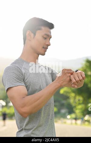 Uomo asiatico sportivo e bello in un abbigliamento sportivo che guarda il suo smartwatch sportivo, controllando il polso o la frequenza cardiaca. Foto Stock