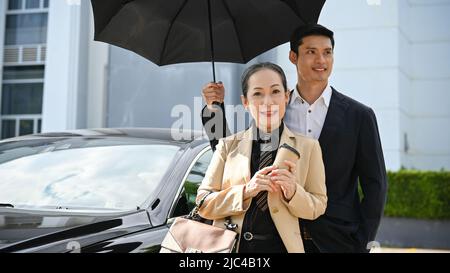 Donna d'affari asiatica invecchiata di successo o direttore esecutivo in piedi accanto alla sua auto di lusso con il bodyguard maschio bello sotto l'ombrello. Foto Stock
