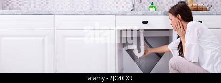 Vista posteriore di una donna che cerca di fermare la perdita di acqua dal tubo di lavello in cucina Foto Stock