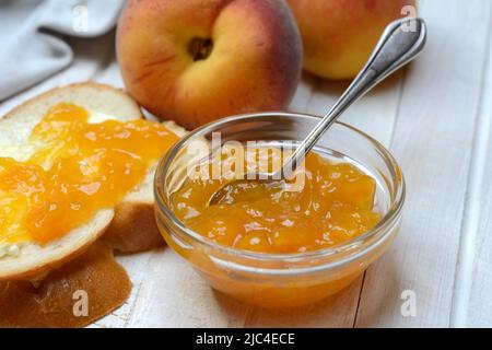 Confettura di pesche in un recipiente di vetro con cucchiaio, fetta di pane e pesche Foto Stock